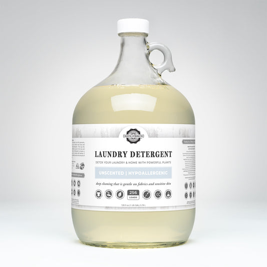 A large, clear glass jug of Rustic Strength Laundry Detergent | Unscented features a white cap and handle. The white label highlights “detox your laundry,” “256 loads,” and “powered by plants” in this biodegradable formula.