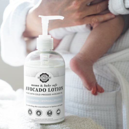 A bottle of Rustic Strength Avocado Lotion | Mama + Baby Safe, made with plant-based ingredients and labeled hypoallergenic & unscented, stands in focus as an adult in a white robe holds a baby, the babys foot visible in the background.