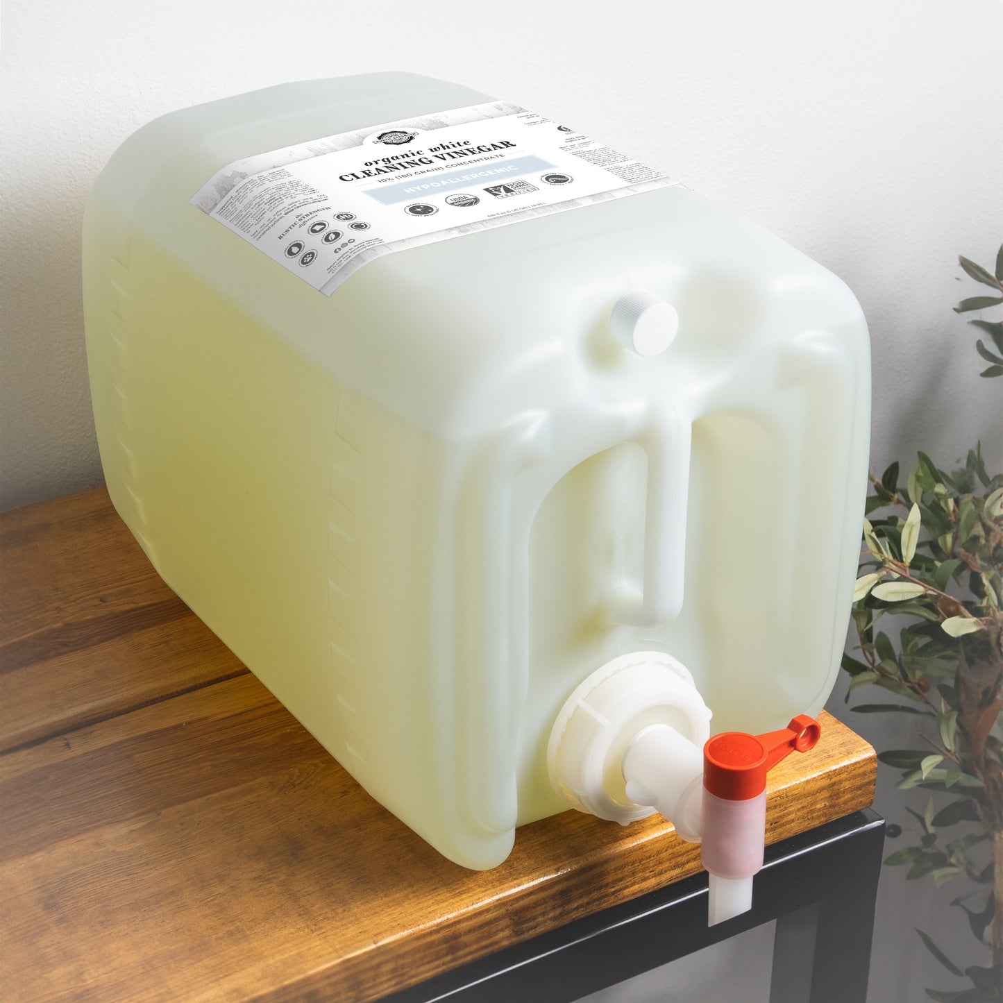A large translucent container of Rustic Strength Organic White Cleaning Vinegar | 5% (50 grain) with a white label and red-tipped spigot sits on wood beside a leafy plant. This biodegradable formula is an eco-friendly cleaning solution.