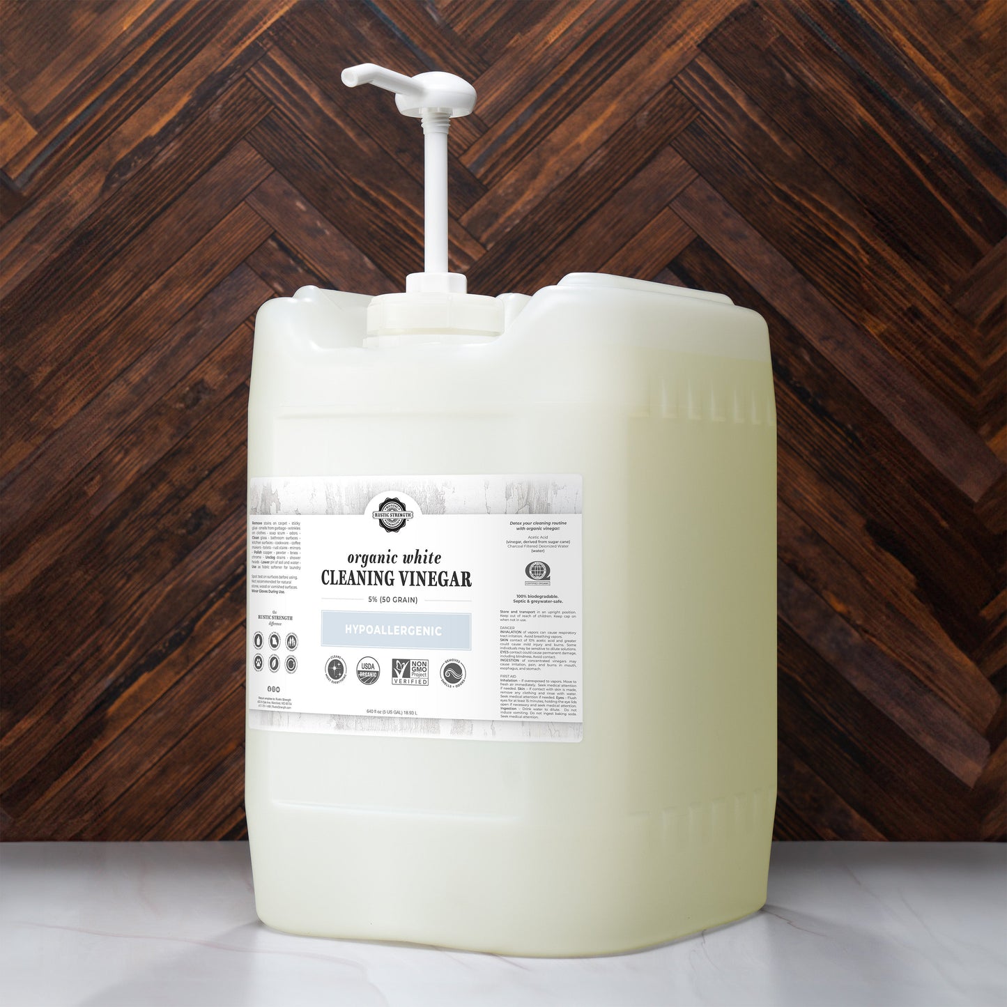 A large biodegradable plastic container with a pump, labeled Rustic Strength Organic White Cleaning Vinegar | 5% (50 grain), is placed on a wooden surface in front of a chevron-patterned wood wall.