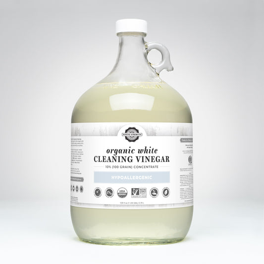 A large clear glass jug of Rustic Strength Organic White Cleaning Vinegar Concentrate | 10% (100 grain), with a white and gray label noting its hypoallergenic, biodegradable formula, is displayed against a plain white background.
