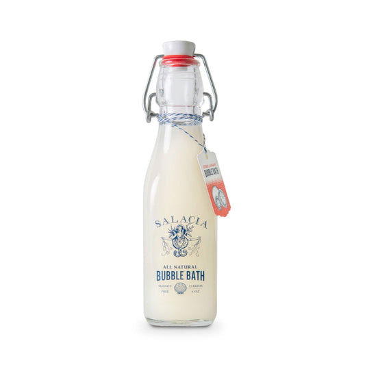 A clear glass bottle with a swing-top lid contains a creamy, moisturizing liquid labeled Salacia Salts Citrus & Sunrays Bubble Bath. A tag is tied around the neck of the bottle with blue and white string.