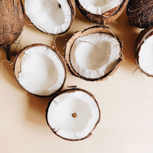 Several brown coconuts are halved and arranged on a light beige surface, revealing their white flesh and rough shells. Some empty halves evoke the luxurious, coconut oil-infused moisturizing benefits of Salacia Salts Coconut Crush Bubble Bath. Fibers are visible.