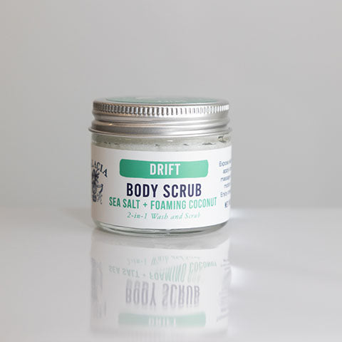 A small glass jar with a silver lid labeled "Drift Sea Salt & Foaming Body Scrub" by Salacia Salts, a 2-in-1 exfoliating wash and scrub, sits on a glossy white surface against a plain background.