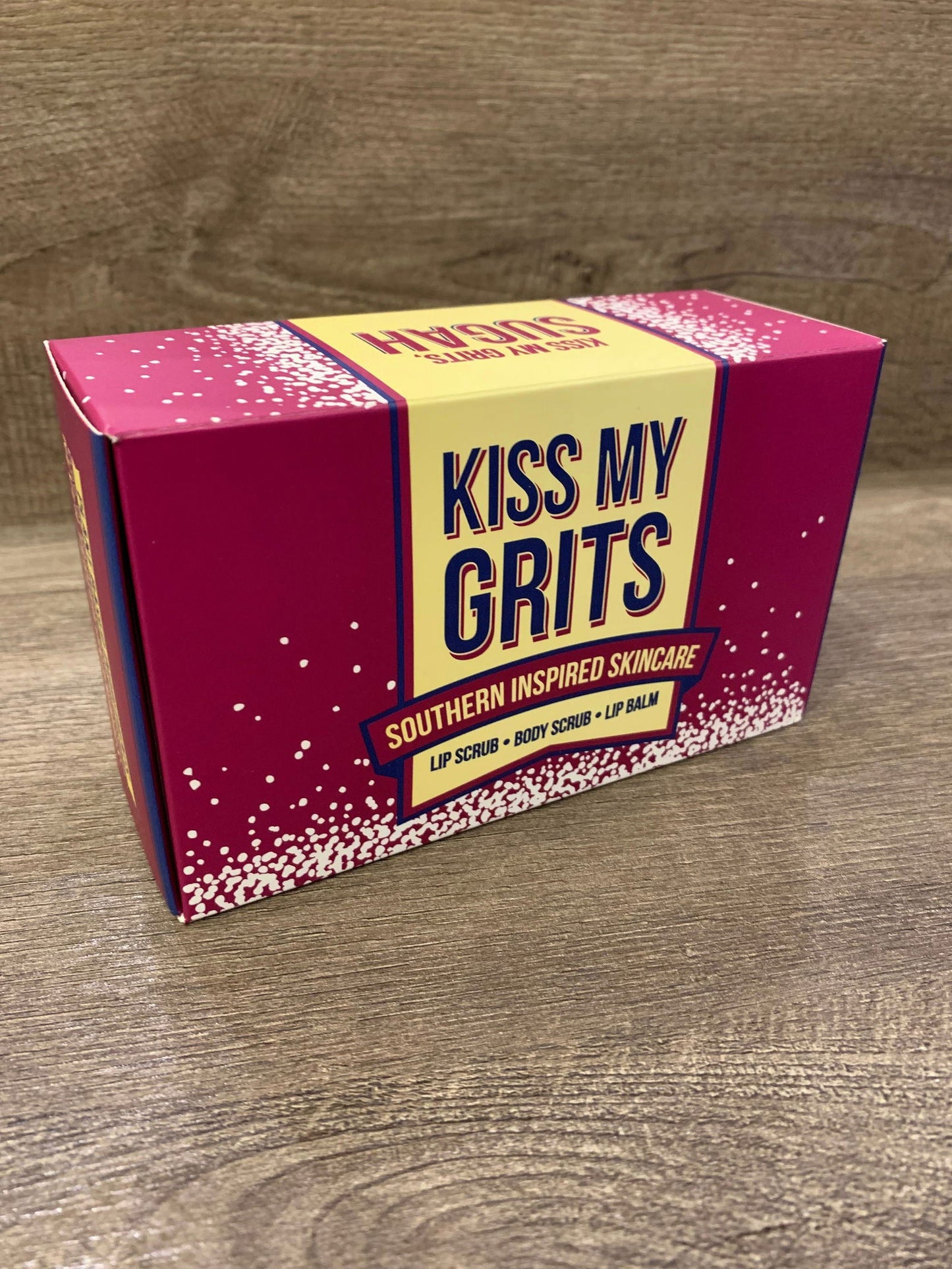A rectangular Kiss My Grits Gift Box by Salacia Salts rests on a wooden surface. The pink and yellow speckled box features Lemon Sugah! scented body scrub, lip scrub, and classic lip balm.