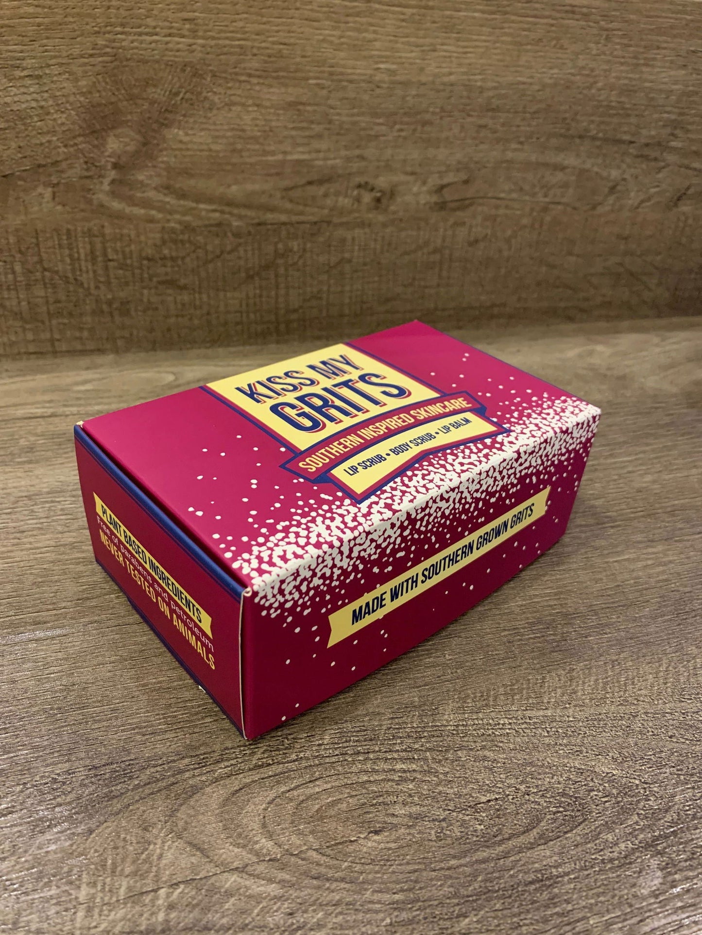 A magenta Kiss My Grits Gift Box by Salacia Salts rests on wood, paired with a classic lip balm. The box reads “South Carolina Stone Ground Grits” and “Made with Southern Grown Grits” in yellow and white lettering.