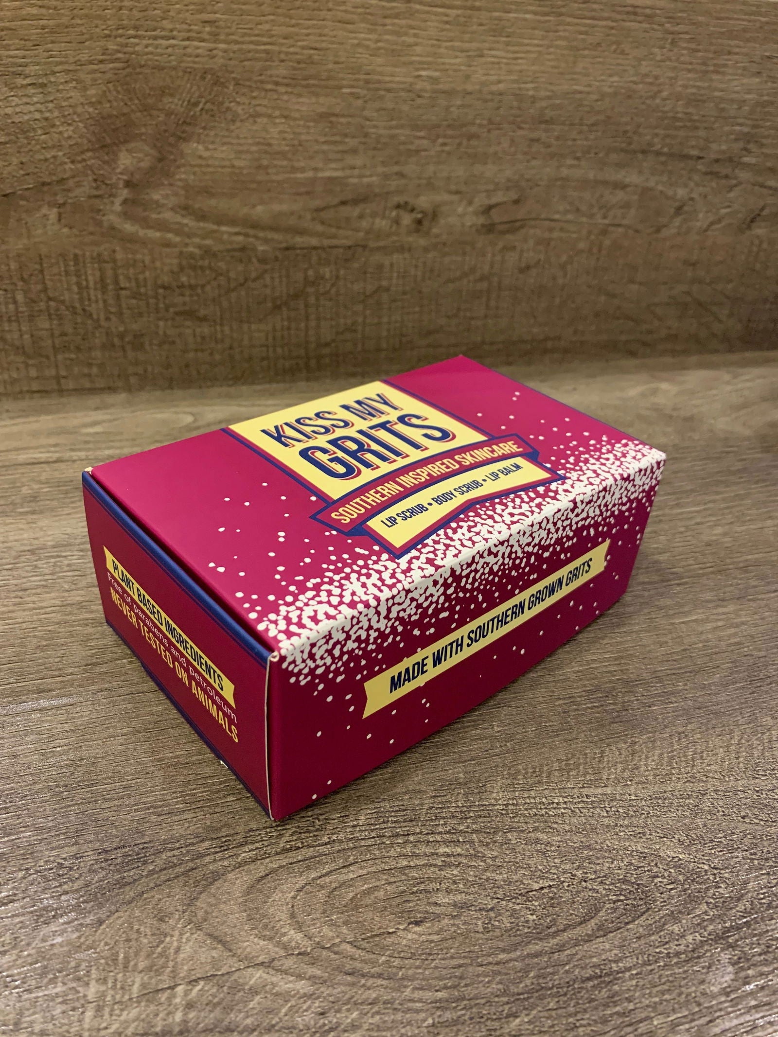 A magenta Kiss My Grits Gift Box by Salacia Salts rests on wood, paired with a classic lip balm. The box reads “South Carolina Stone Ground Grits” and “Made with Southern Grown Grits” in yellow and white lettering.