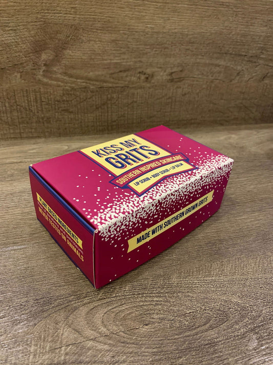 A magenta Kiss My Grits Gift Box by Salacia Salts rests on wood, paired with a classic lip balm. The box reads “South Carolina Stone Ground Grits” and “Made with Southern Grown Grits” in yellow and white lettering.
