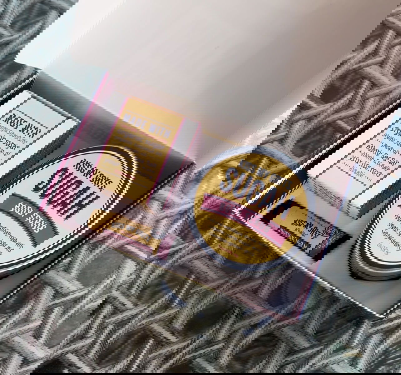 Blurry image of Salacia Salts' Kiss My Grits Gift Box, featuring several cosmetic product boxes including a classic lip balm and Lemon Sugah! Scented Body Scrub, on a woven surface. Some text and logos are visible but unclear.