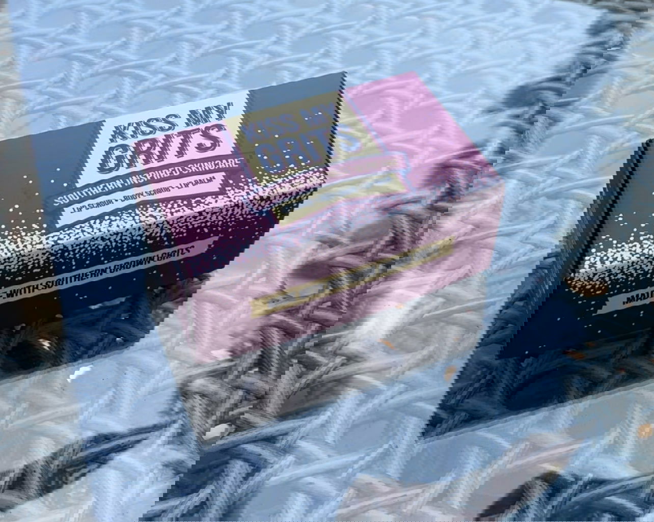 Two black Kiss My Grits Gift Boxes from Salacia Salts sit on a textured silver metal surface. A pink lip balm is nestled between the boxes, partially obscuring the text below each label.