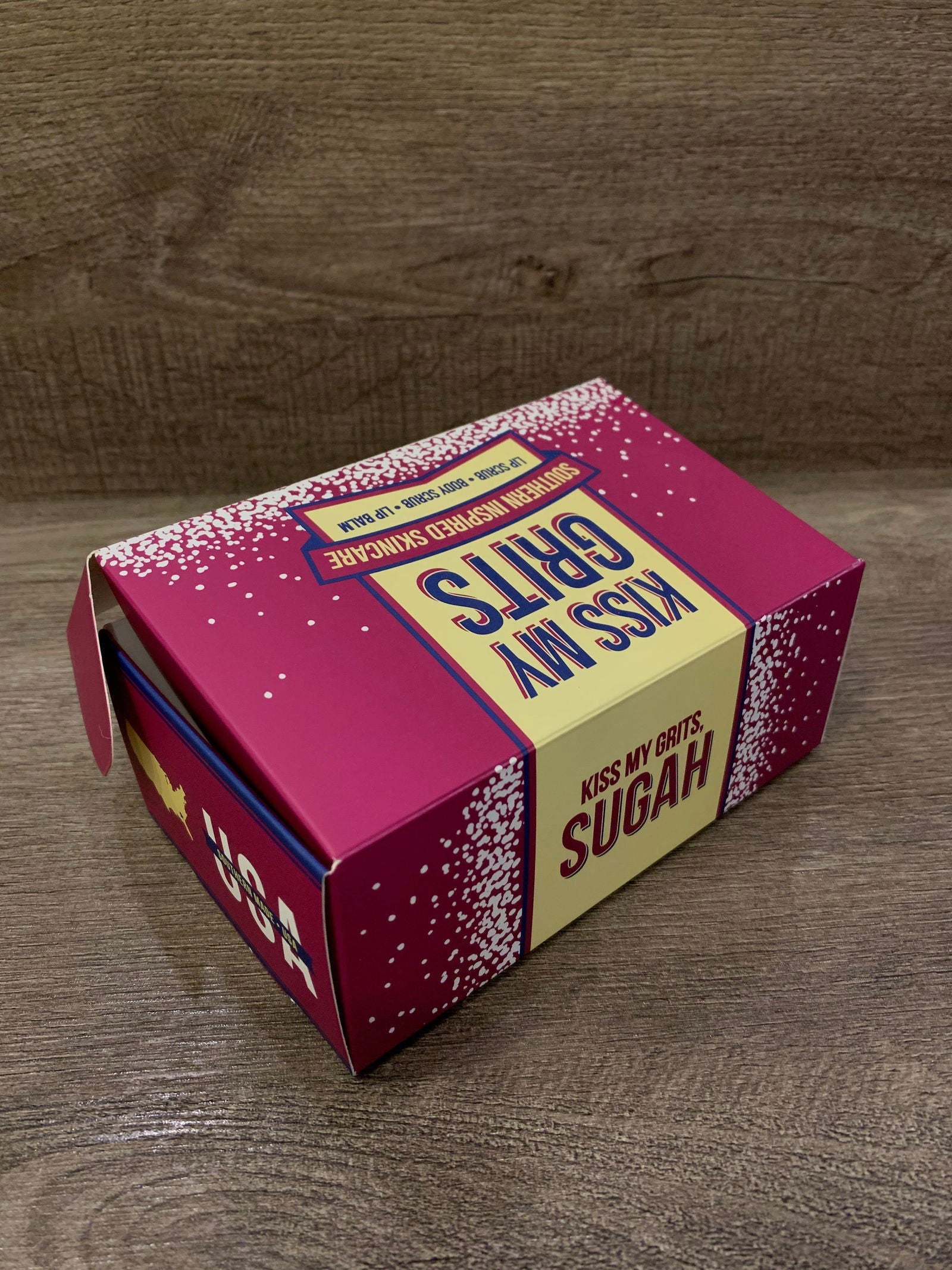 A pink and yellow Kiss My Grits Gift Box by Salacia Salts sits on a wooden surface. The top features bold blue lettering and a sprinkle design—ideal packaging for their classic lip balm or Lemon Sugah! Scented Body Scrub.