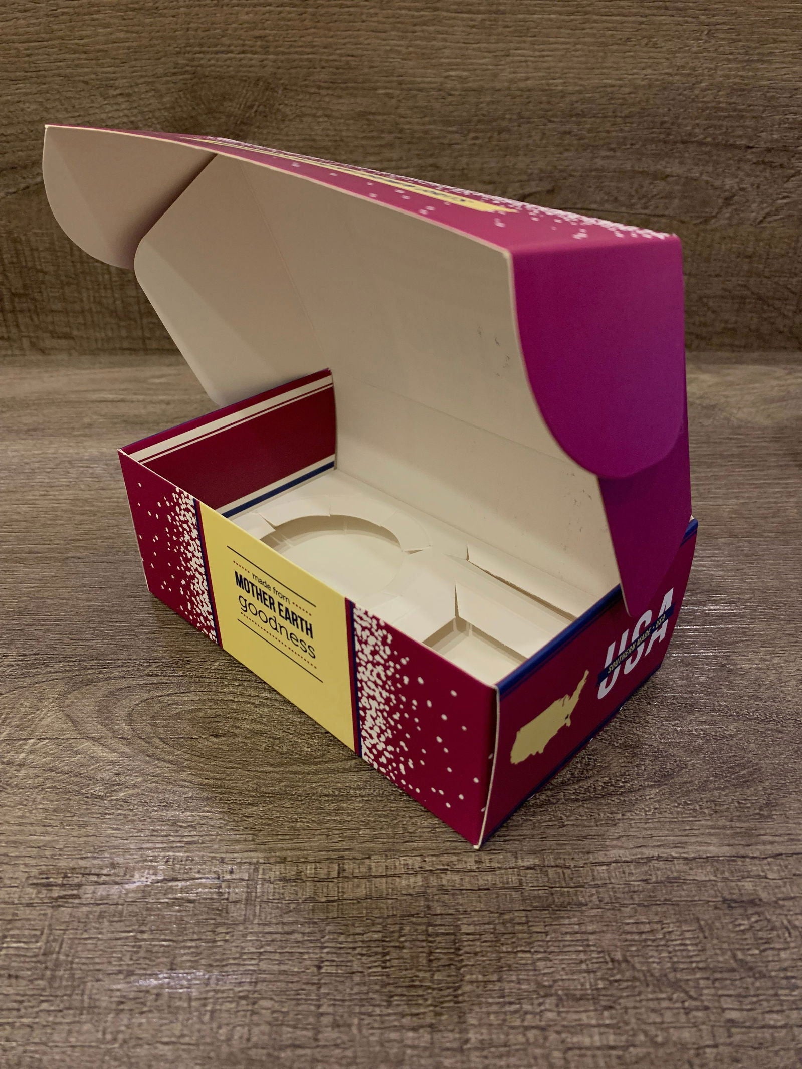 A vibrant, open Kiss My Grits Gift Box by Salacia Salts rests on a wooden surface, featuring red, pink, and yellow sides with the brand name and product details visible on the outside.