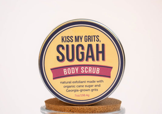 A round tin of Salacia Salts Kiss My Grits Sugah! Body Scrub, an exfoliating sugar scrub with organic cane sugar and Georgia-grown grits, rests on a cork lid against a light background.