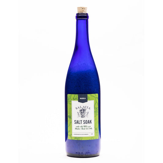 A tall, deep blue glass bottle with a cork, labeled “Magnolia Upcycled Salt Soak” by Salacia Salts, contains magnolia-scented bath salts—an ideal luxury gift. The bottle is shown upright against a plain white background.