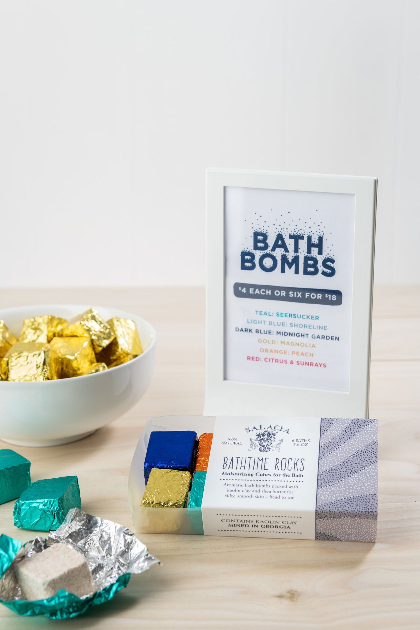 A display of Salacia Salts’ Midnight Garden BathTime Rocks (Set of 6): a framed info sign, a bowl of gold and green wrapped cubes, and a clear box—perfect for an essential oils-infused, moisturizing bath soak.