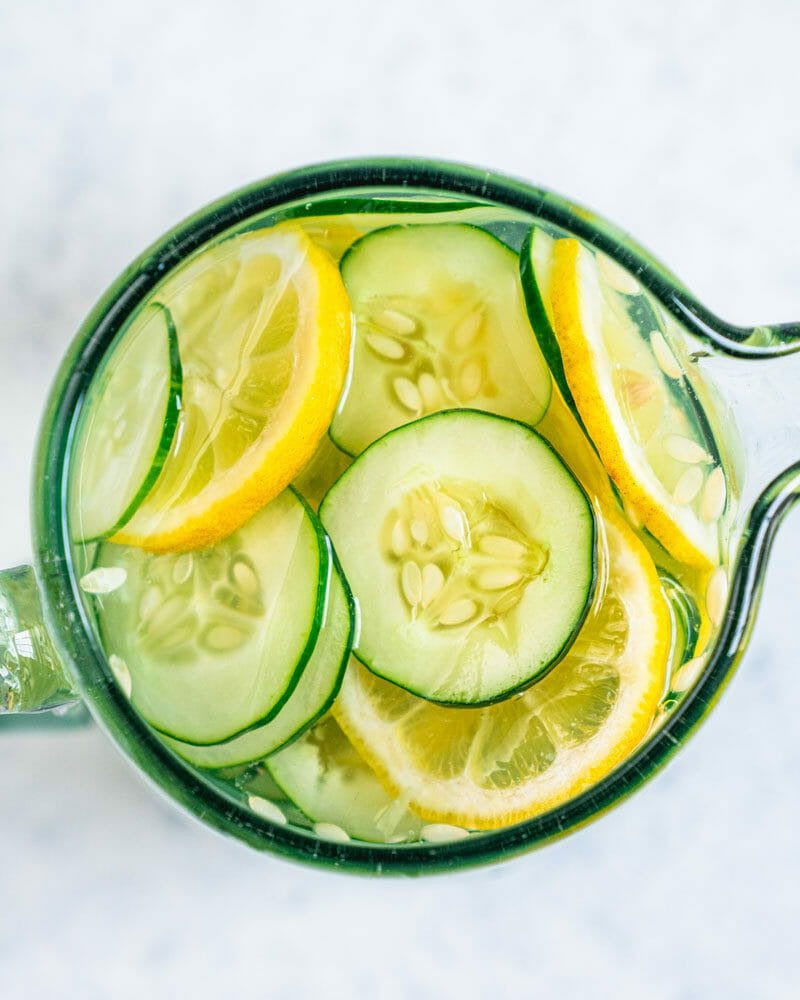 A glass pitcher with water, cucumber, and lemon slices viewed from above on a light surface. The fresh aroma recalls the soothing soak of Salacia Salts Midnight Garden BathTime Rocks (Set of 6) with essential oils.