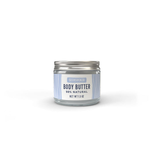 A clear glass jar with a silver lid, labeled Salacia Salts Seersucker Body Butter, 98% natural, Net Wt 1.5 oz, features light blue and white stripes and a clean cotton-scented body butter formula.
