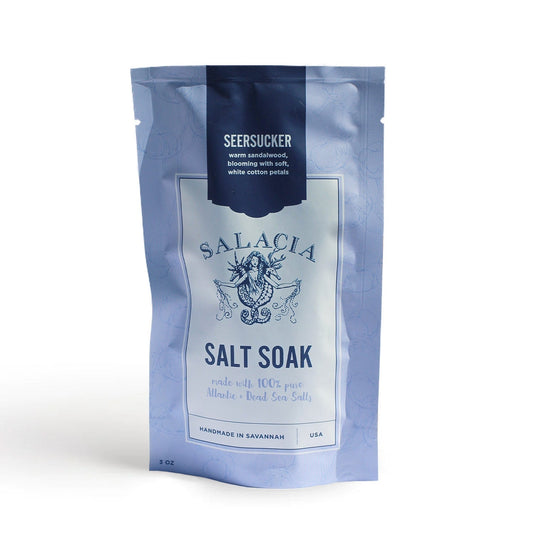 A light blue resealable pouch labeled Salacia Salts Seersucker Small Salt Soak displays decorative art, mentions Atlantic and Dead Sea salts, and lists the scent as sandalwood, cotton petals, and fresh island fragrances.