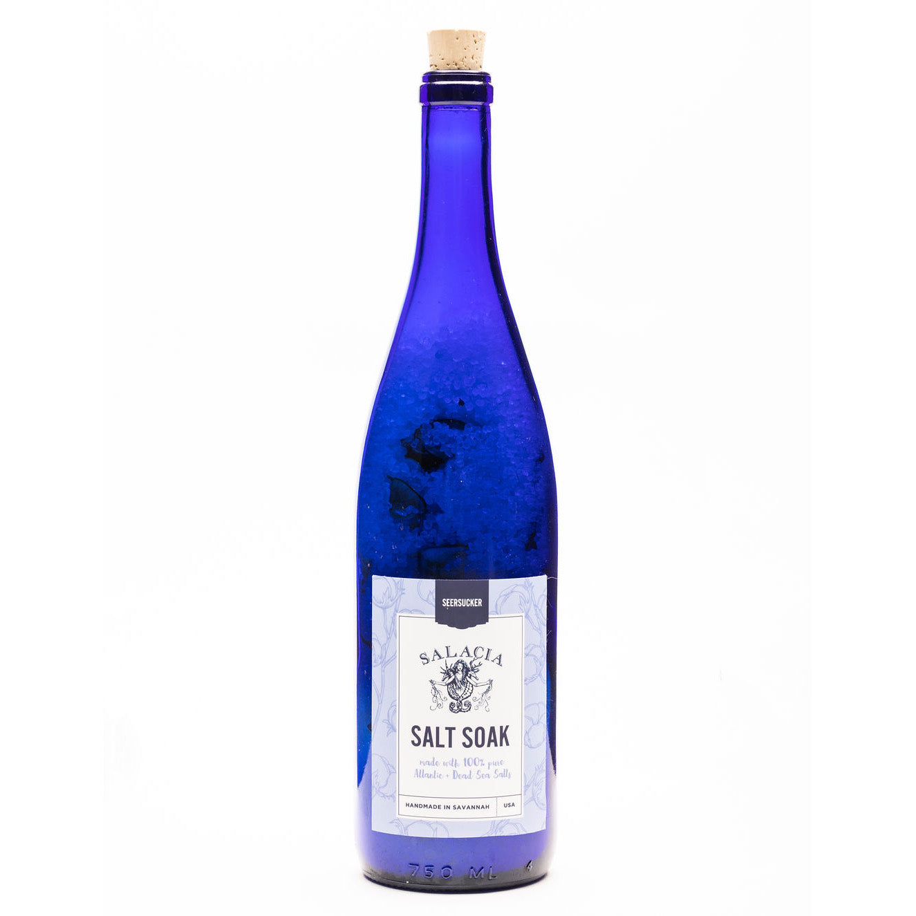 A tall, blue glass bottle with a cork top labeled Seersucker Upcycled Salt Soak by Salacia Salts on a white background. The label describes it as a blend of sea salt with lavender and island fragrances for a soothing, balanced scent.