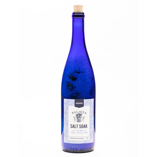 A tall, blue glass bottle with a cork top labeled Seersucker Upcycled Salt Soak by Salacia Salts on a white background. The label describes it as a blend of sea salt with lavender and island fragrances for a soothing, balanced scent.