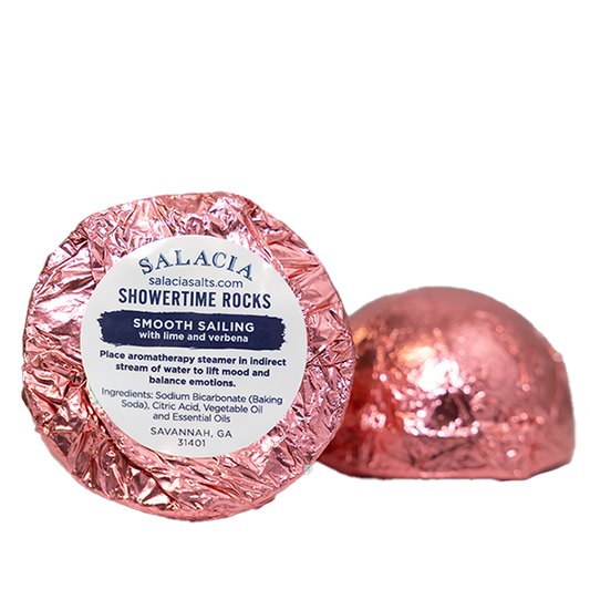 Two round ShowerTime Rocks - Smooth Sailing shower steamers by Salacia Salts, wrapped in shiny pink foil; one has a label with lime and verbena aromatherapy, shown with instructions and ingredients.
