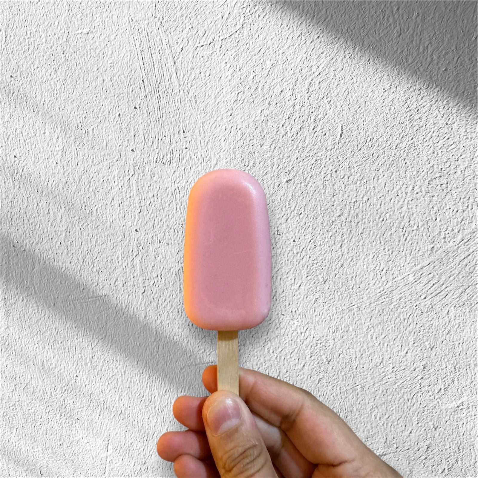 A hand holds Salacia Salts’ Popcycle Lotion Bar for hair, face, or body—reminiscent of summer’s delight and Georgia Peach fragrance—set against a textured white wall with diagonal shadows.