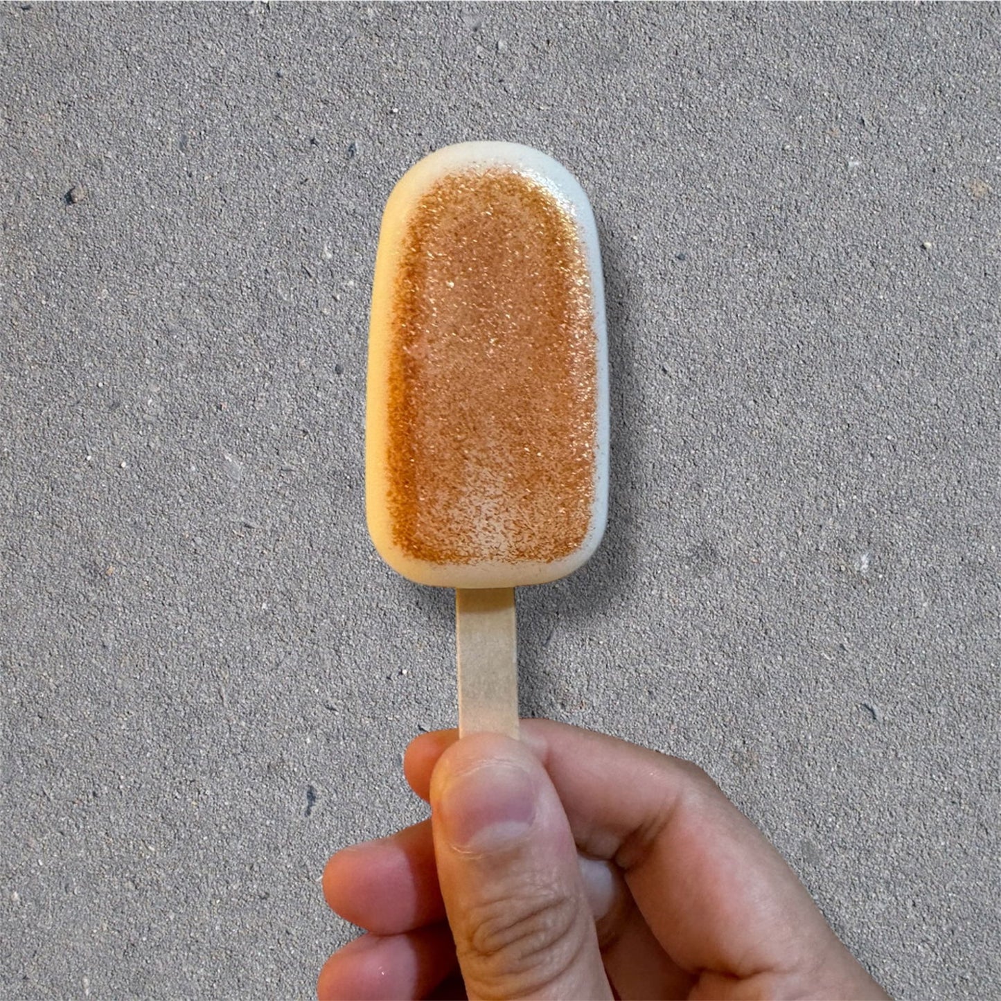 A hand holds Salacia Salts’ Popcycle Lotion Bar for hair, face, or body—its tan, sandy texture on a stick resembles a popsicle, shown against a gray concrete-like background. The bar mimics the look of a sandy patch.