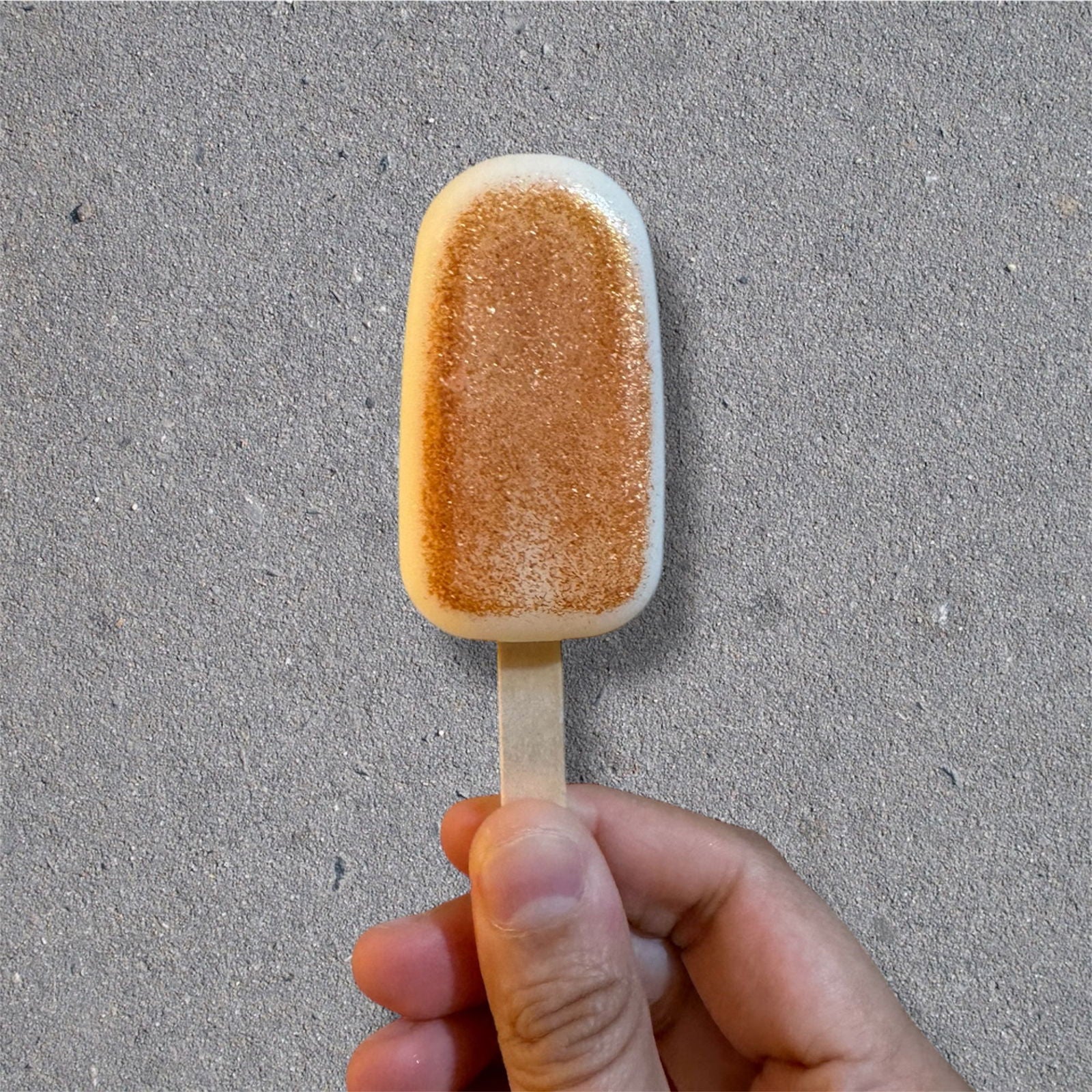A hand holds Salacia Salts’ Popcycle Lotion Bar for hair, face, or body—its tan, sandy texture on a stick resembles a popsicle, shown against a gray concrete-like background. The bar mimics the look of a sandy patch.