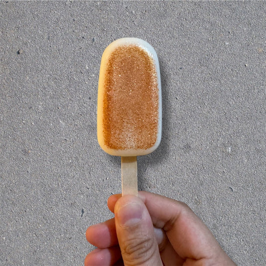 A hand holds Salacia Salts’ Popcycle Lotion Bar for hair, face, or body—its tan, sandy texture on a stick resembles a popsicle, shown against a gray concrete-like background. The bar mimics the look of a sandy patch.