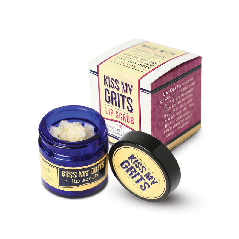 An open small blue jar of Salacia Salts Kiss My Grits Lip Scrub sits with its lid beside it. Behind it, a matching box showcases this natural exfoliating lip treatment with bold yellow, pink, and purple branding.