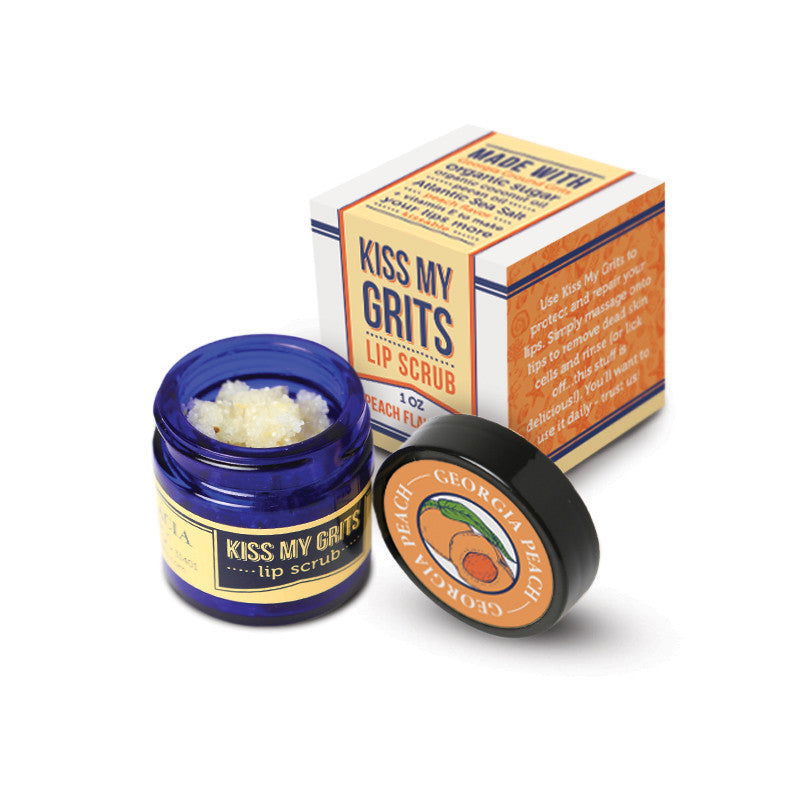 A small blue jar of Salacia Salts Kiss My Grits Lip Scrub sits open, revealing its coarse natural peach scrub. The labeled black lid and product box are shown nearby.