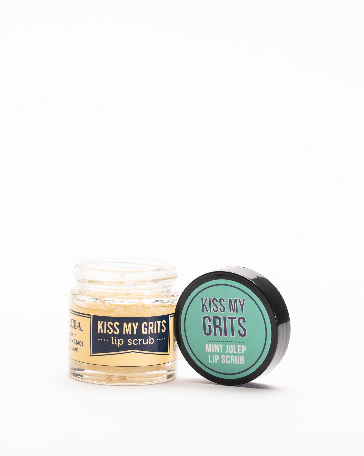 A small open jar of Salacia Salts Kiss My Grits Lip Scrub sits beside its black lid labeled Mint Julep Lip Scrub. The jar contains a pale yellow, natural exfoliating lip treatment on a light background.