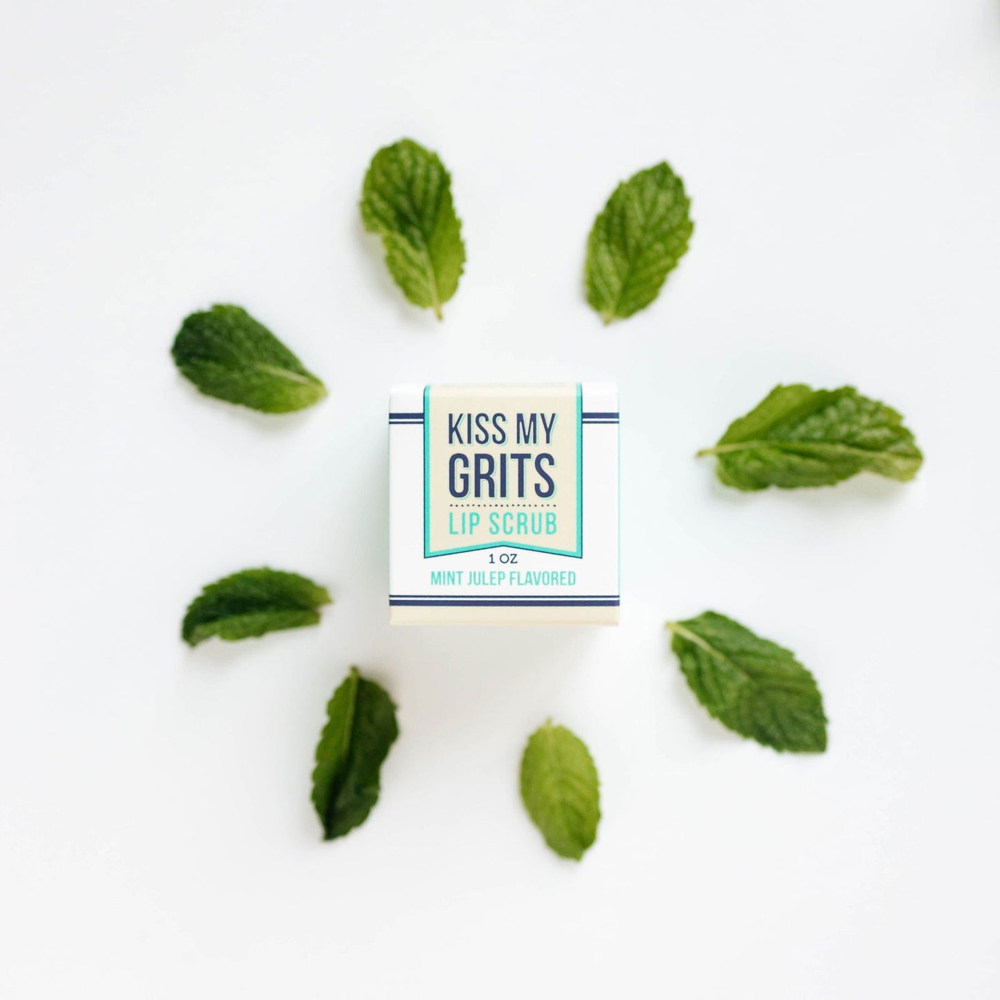 A box of Salacia Salts Kiss My Grits Lip Scrub sits on a white background, encircled by eight mint leaves. This natural exfoliating lip scrub delights with a refreshing mint julep flavor for smooth, soft lips.
