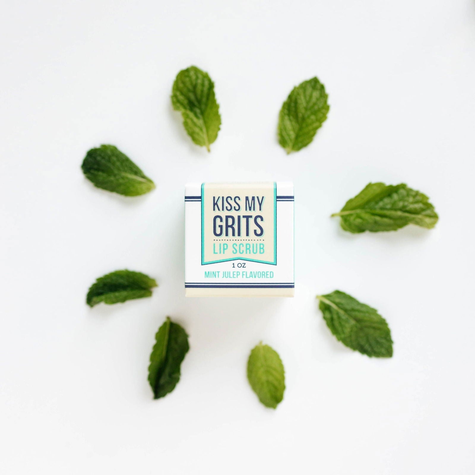 A box of Salacia Salts Kiss My Grits Lip Scrub sits on a white background, encircled by eight mint leaves. This natural exfoliating lip scrub delights with a refreshing mint julep flavor for smooth, soft lips.
