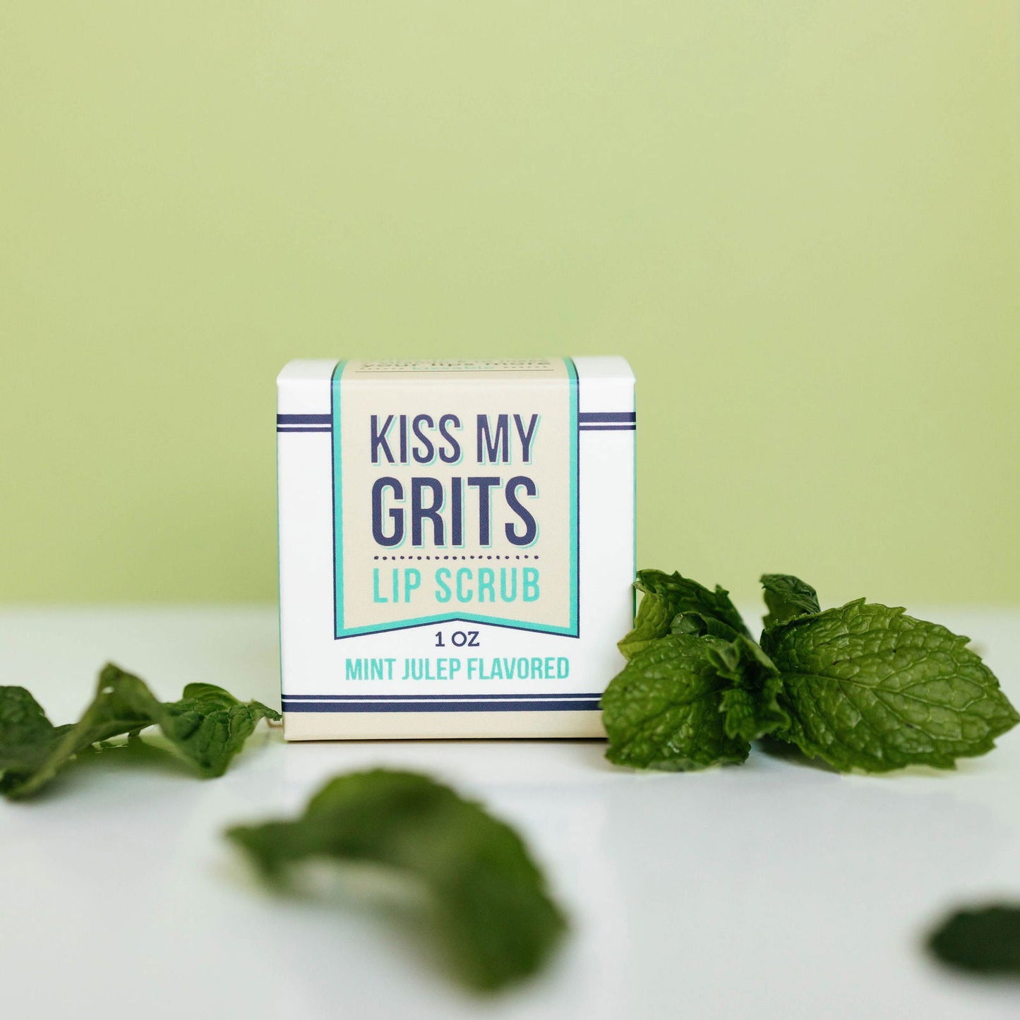 A small box of Salacia Salts Kiss My Grits Lip Scrub, Mint Julep flavor, sits on a white surface among fresh mint leaves and a pale green backdrop—a refreshing exfoliating lip treatment for naturally soft lips.