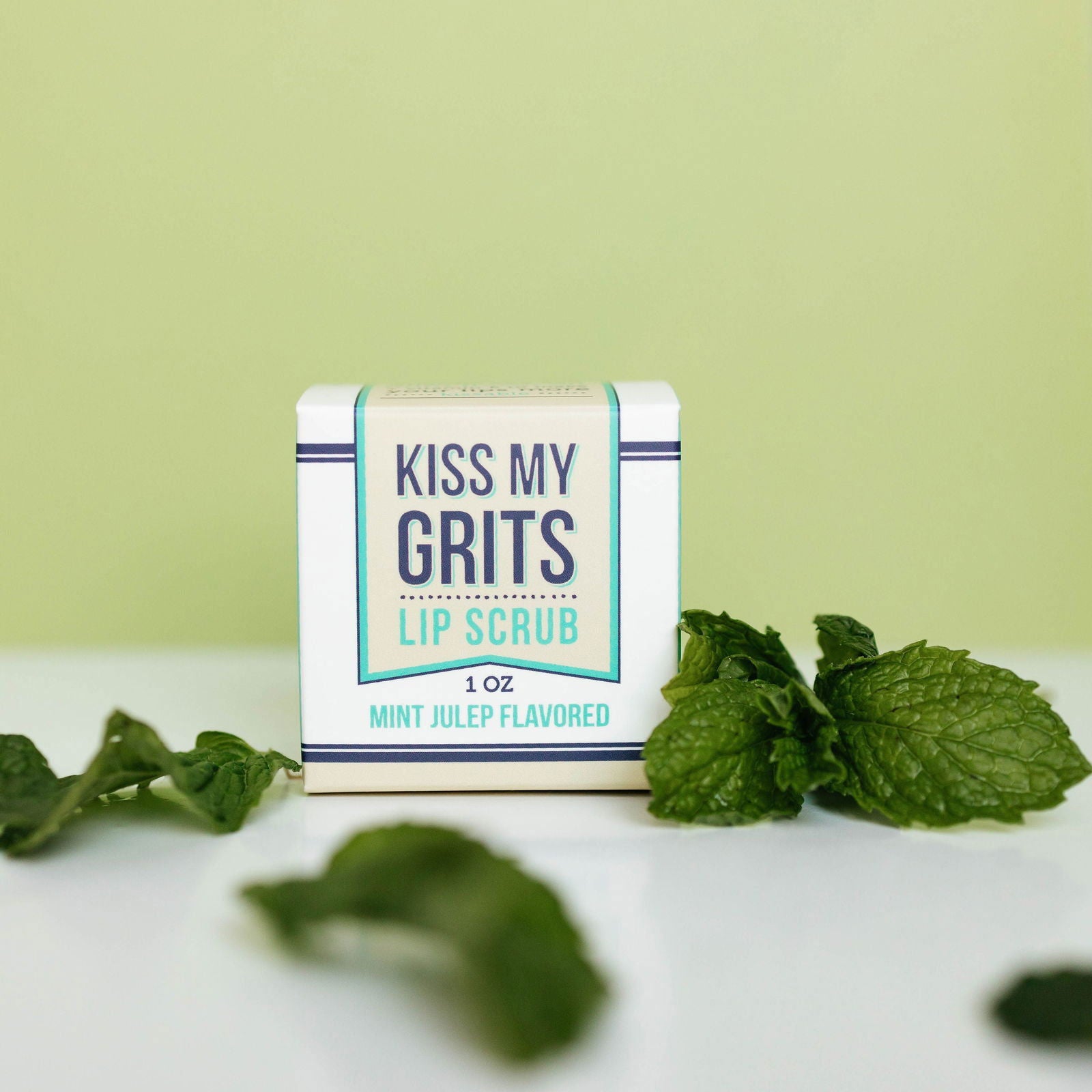 A small box of Salacia Salts Kiss My Grits Lip Scrub, Mint Julep flavor, sits on a white surface among fresh mint leaves and a pale green backdrop—a refreshing exfoliating lip treatment for naturally soft lips.