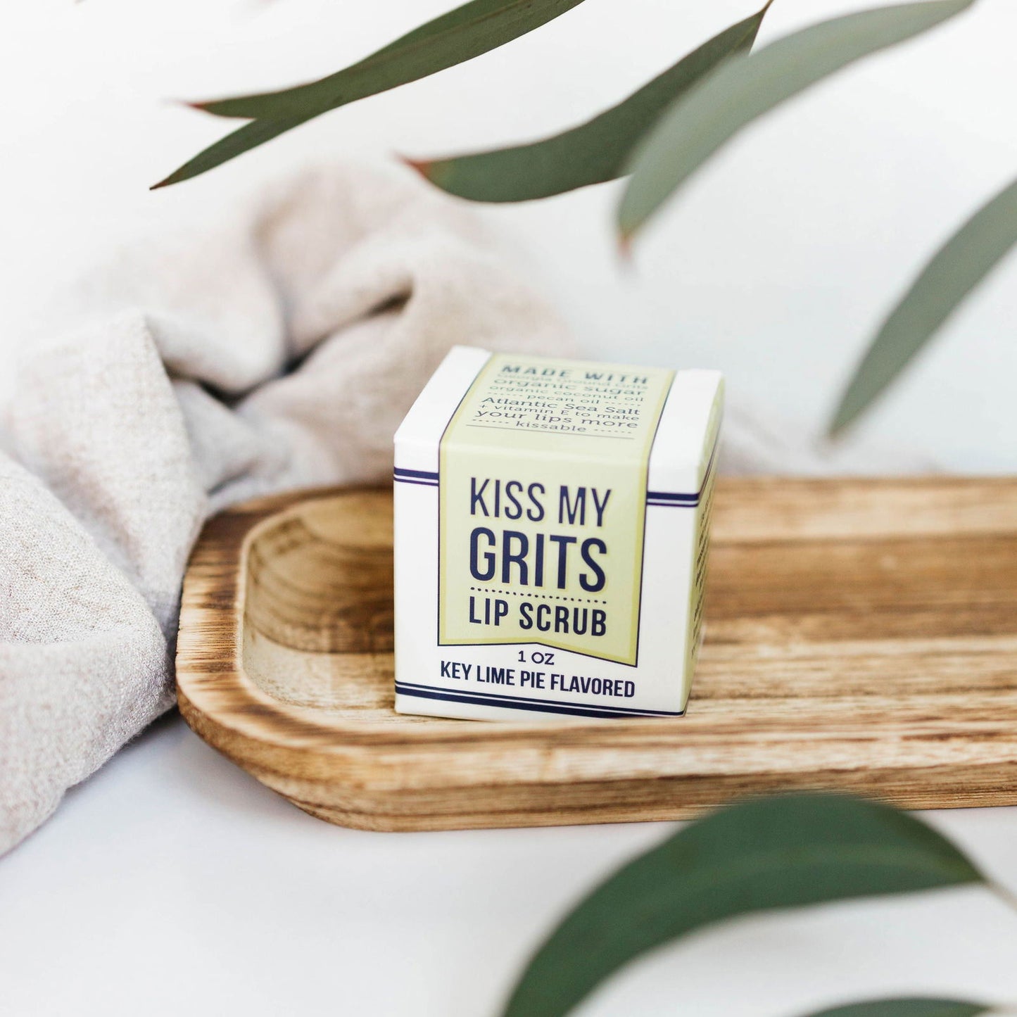 A box labeled Kiss My Grits Lip Scrub by Salacia Salts, 1 oz, Key Lime Pie Flavored, is arranged on a wooden tray with green leaves and fabric—a fresh display for this natural lip scrub.