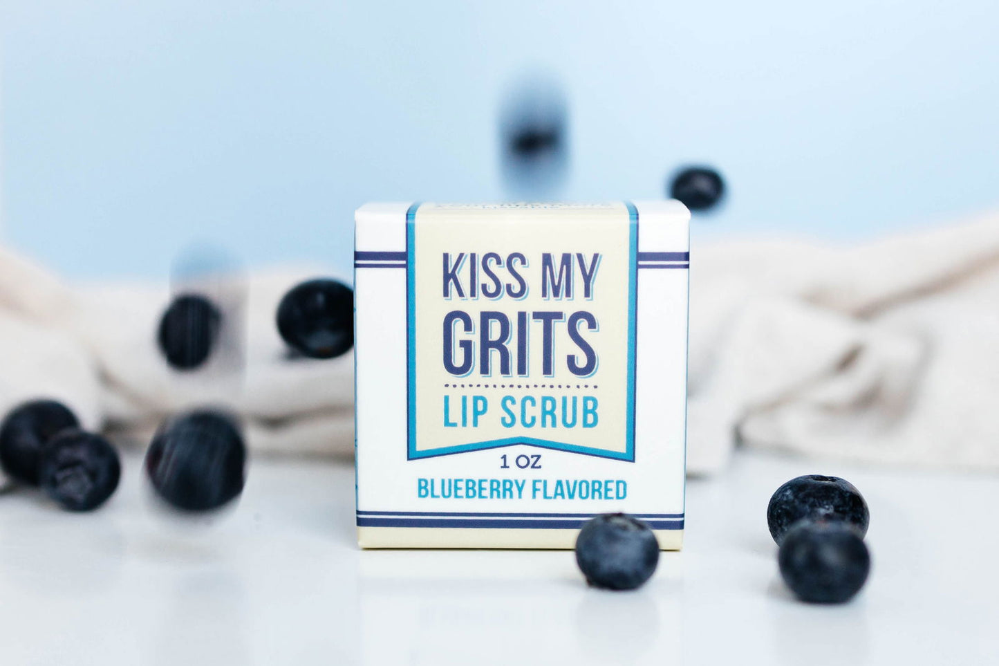 A box of Salacia Salts Kiss My Grits Lip Scrub in blueberry flavor rests on a white surface with scattered blueberries and a neutral background—ideal for a refreshing, natural lip exfoliation.