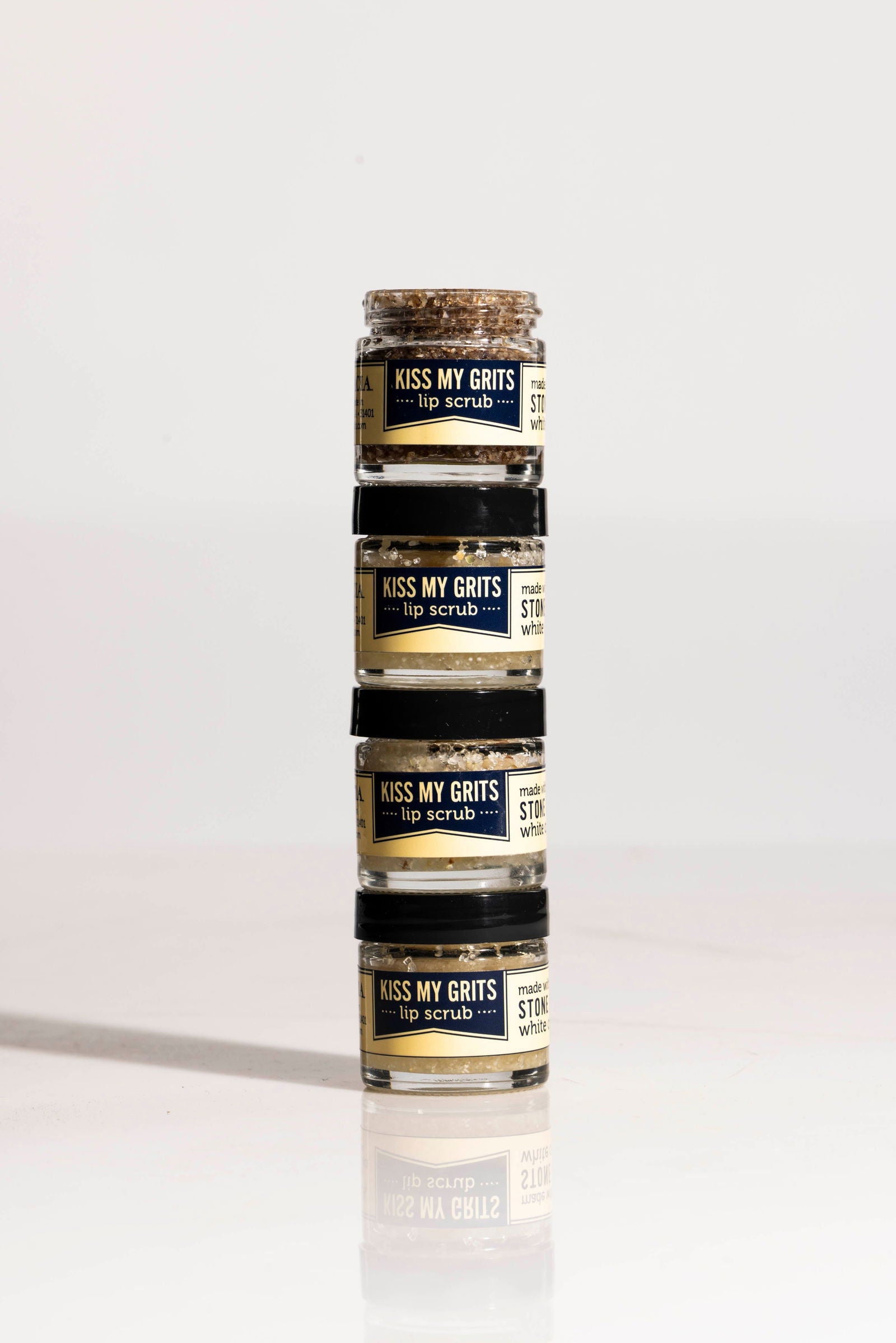 A vertical stack of four small Salacia Salts Kiss My Grits Lip Scrub jars, filled with dark scrub, sits on a shiny surface against a plain white background.
