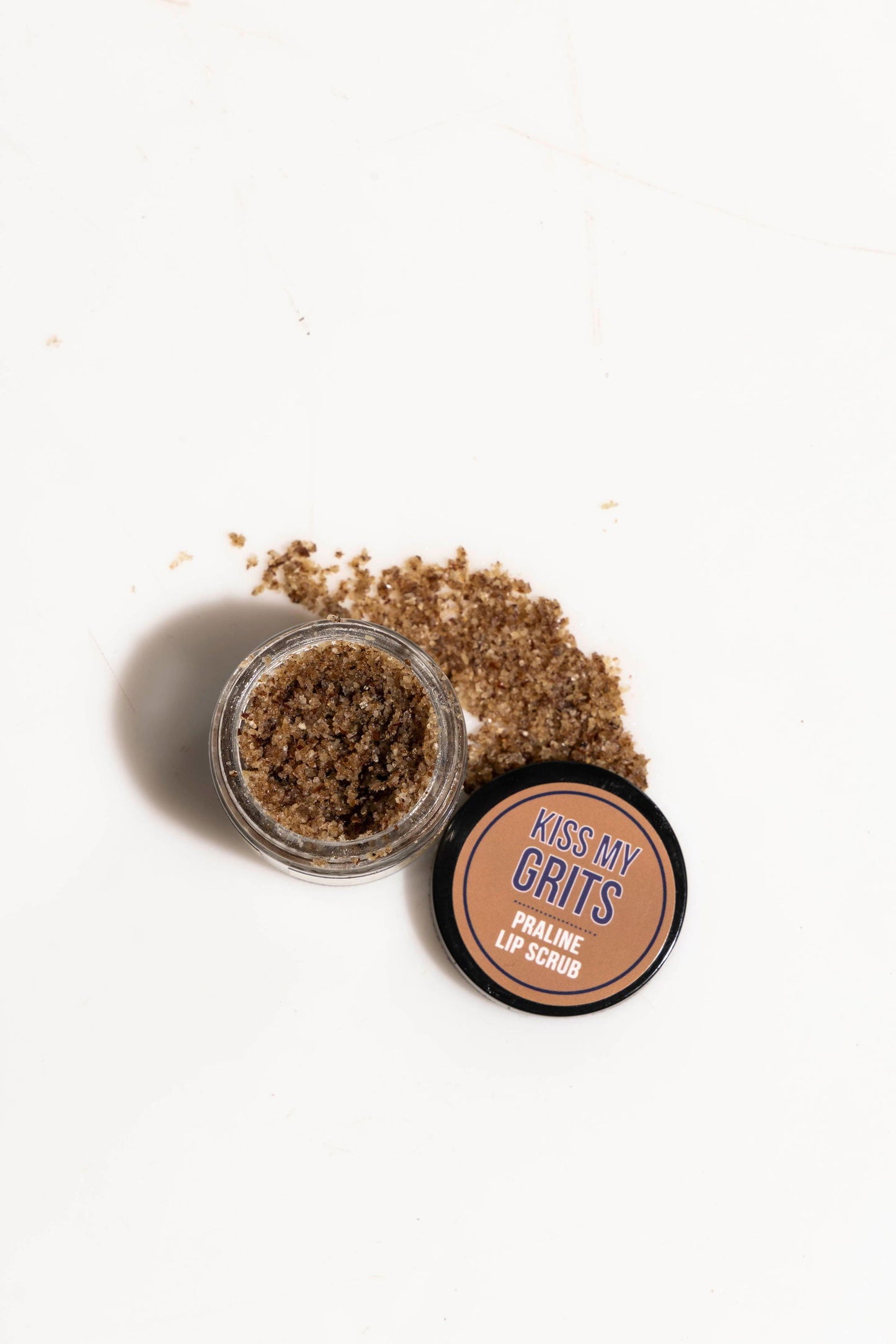 An open container of Salacia Salts Kiss My Grits Lip Scrub spills onto a white surface. The black lid beside it displays the product name in orange and white—an exfoliating treatment for smooth lips.