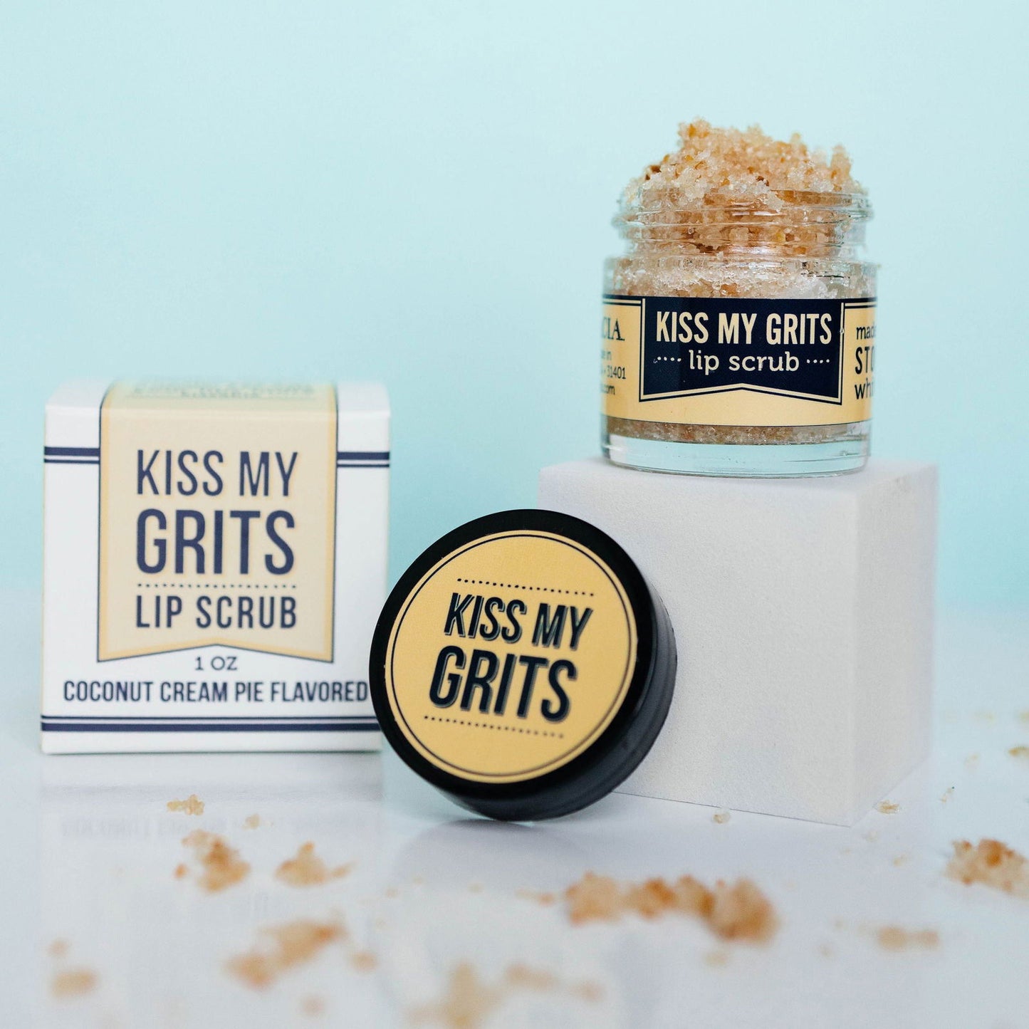An open jar of Salacia Salts Kiss My Grits Lip Scrub sits on a white block with some scrub spilled nearby. The product box and a closed container are also shown, all set against a light blue background.