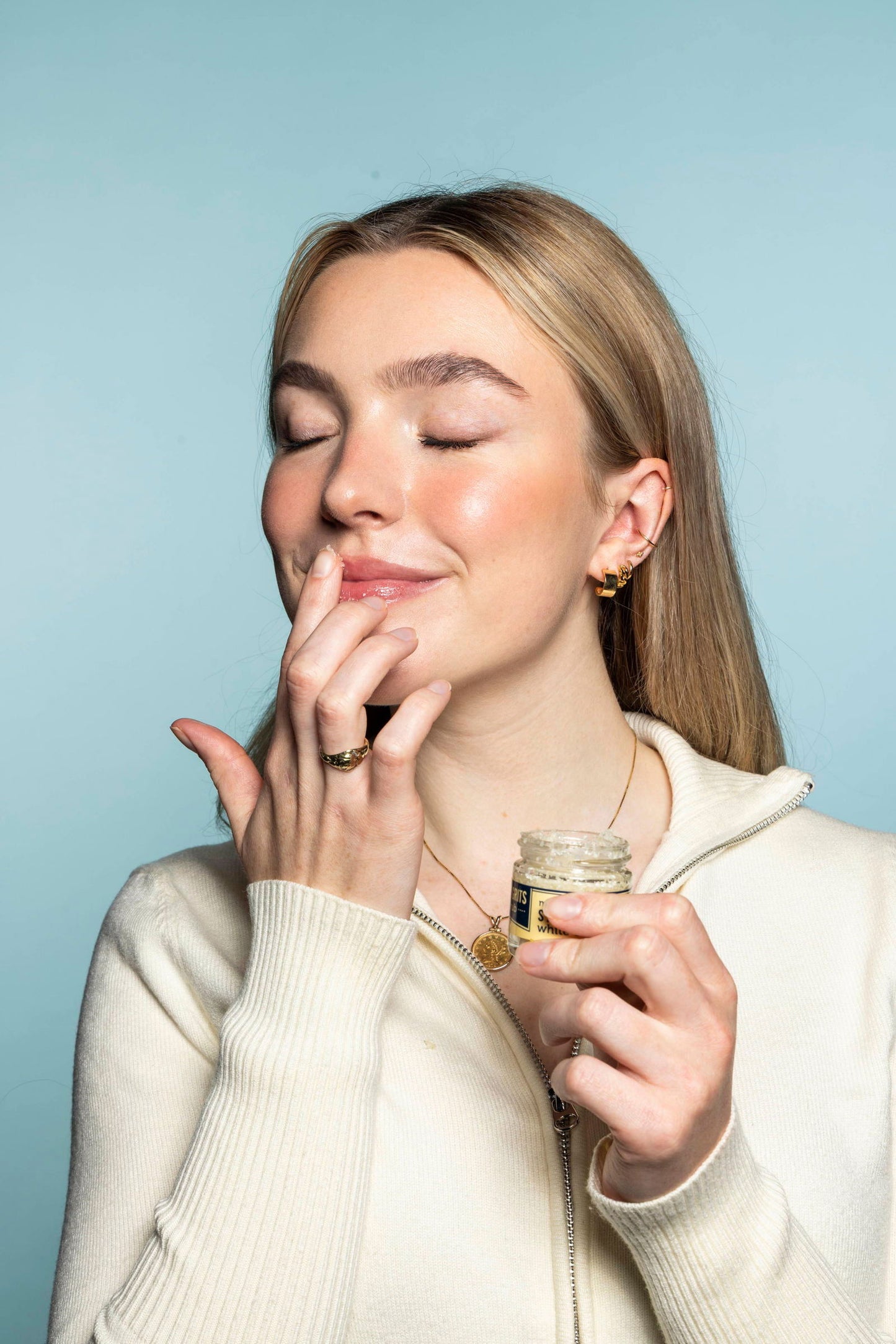 A woman with closed eyes and a slight smile applies Salacia Salts’ Kiss My Grits Lip Scrub to her lips with her finger. She holds the small jar, wearing a cream-colored top against a light blue background.