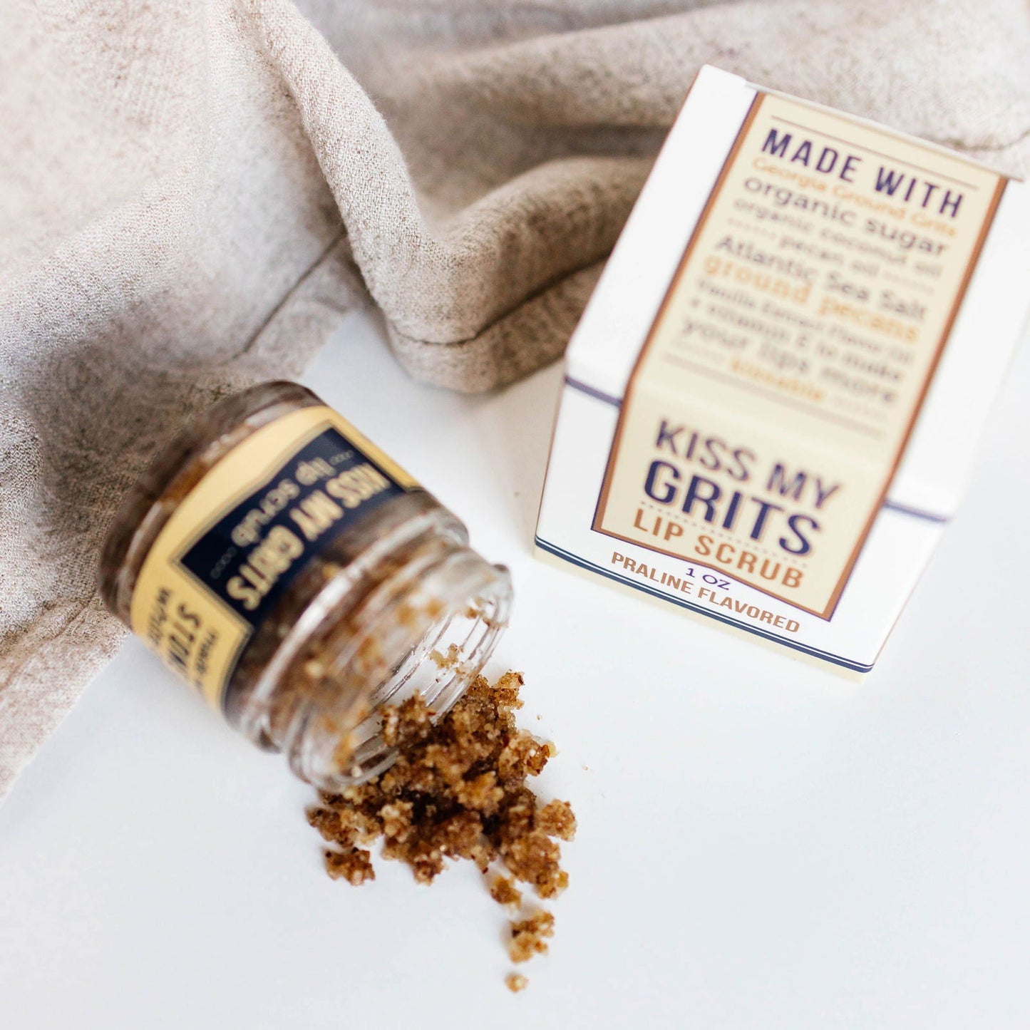 A small glass jar labeled Salacia Salts Kiss My Grits Lip Scrub, an exfoliating lip treatment, is tipped over with brown sugar scrub spilling out. Its box is nearby, and a beige cloth appears in the background.
