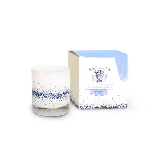 A white hand-poured soy blend candle in a glass holder with blue speckles sits next to its box labeled Salacia Salts Seersucker Signature Candle, featuring a blue and white design and the scent Seiruser.