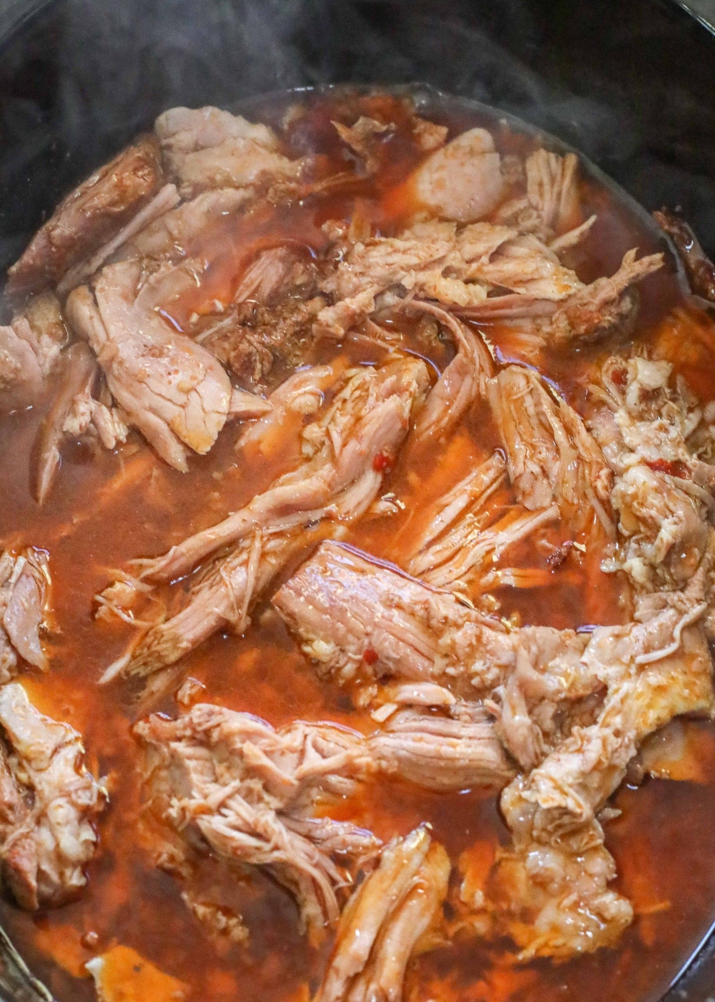 Shredded pork simmers in SavannahSauceCompanys NEW Tangy Orange BBQ Sauce Case, filling the pot with steam and a rich, reddish-brown orange aroma.