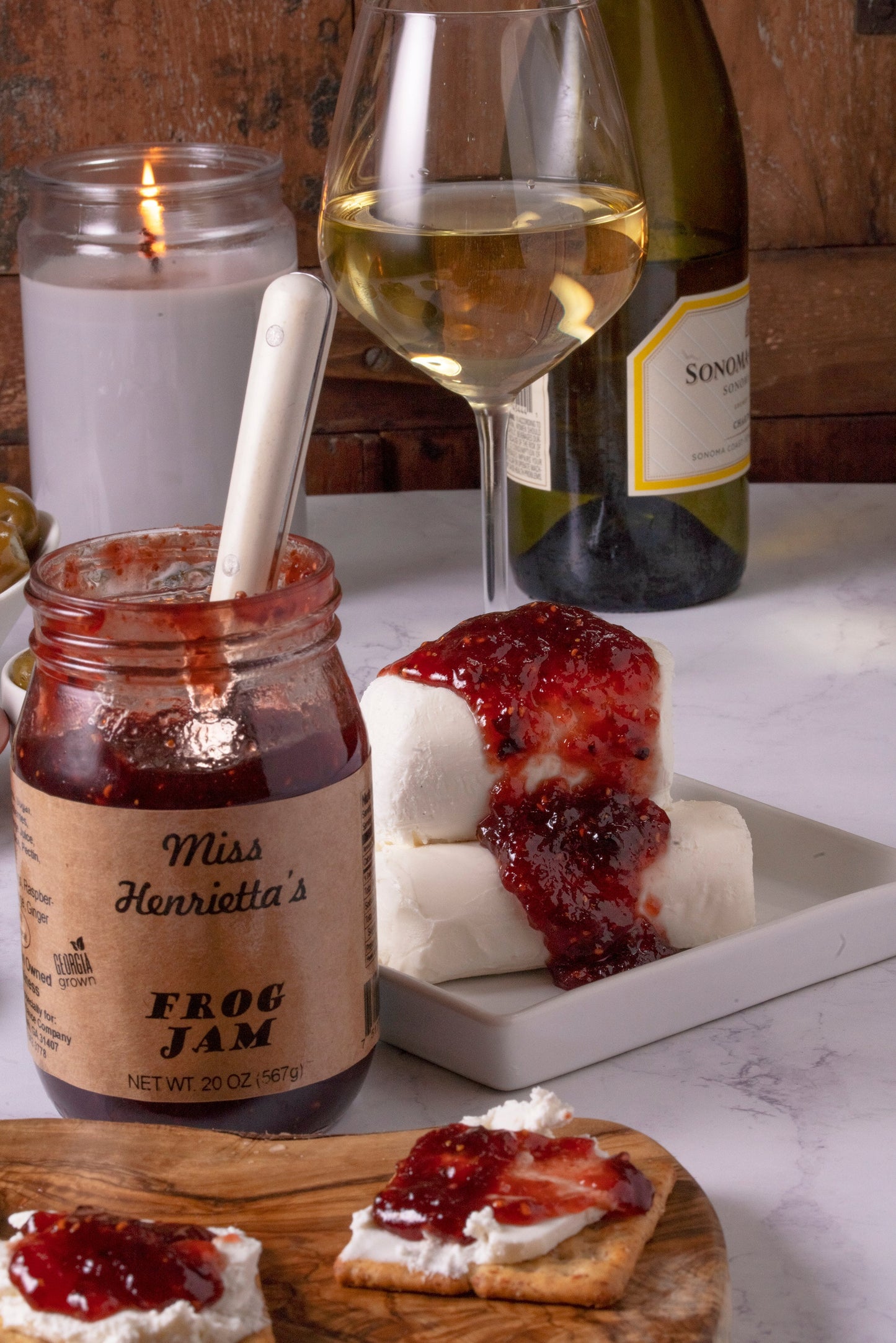 A 12-jar case of SavannahSauceCompany’s FROG Jam sits by cheese topped with jam, a glass of white wine, a lit candle, and a cracker with cheese and jam on marble.