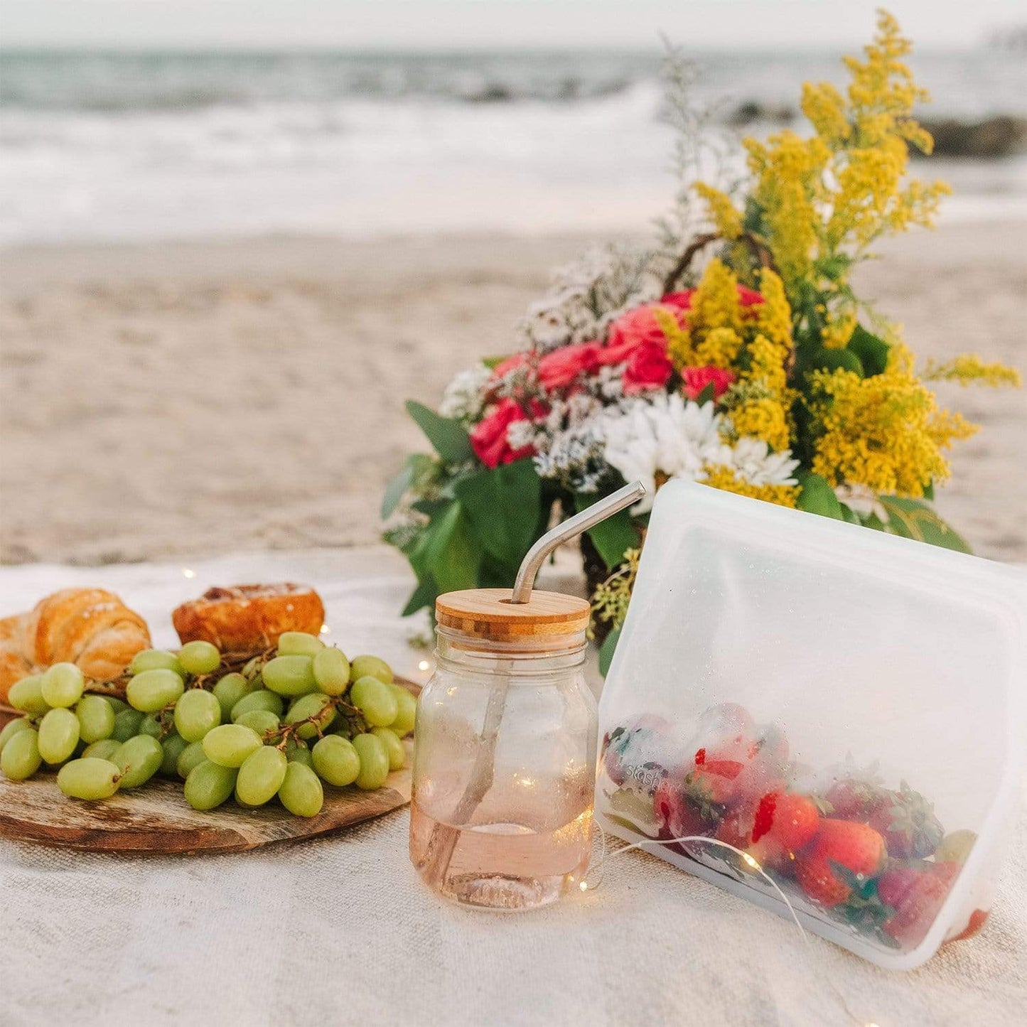A beach picnic with green grapes, pastries, strawberries in a reusable bag, a drink in a jar topped with the Bamboo Mason Jar Lid from ZeroWasteStore.com, and colorful flowers—all set against the stunning ocean backdrop.