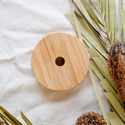 The Bamboo Mason Jar Lid by ZeroWasteStore.com, a round flat bamboo lid with a center hole, rests on white textured fabric and is surrounded by dried green leaves and brown seed pods.