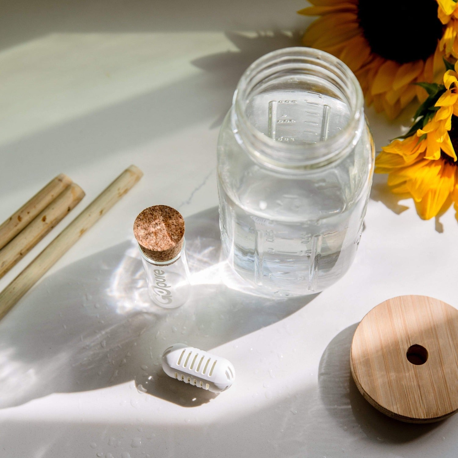 A Bamboo Mason Jar Lid by ZeroWasteStore.com rests on a white surface next to a glass jar of water, cork-topped glass vial, bamboo straws, white filter, and two yellow sunflowers as sunlight casts gentle shadows.