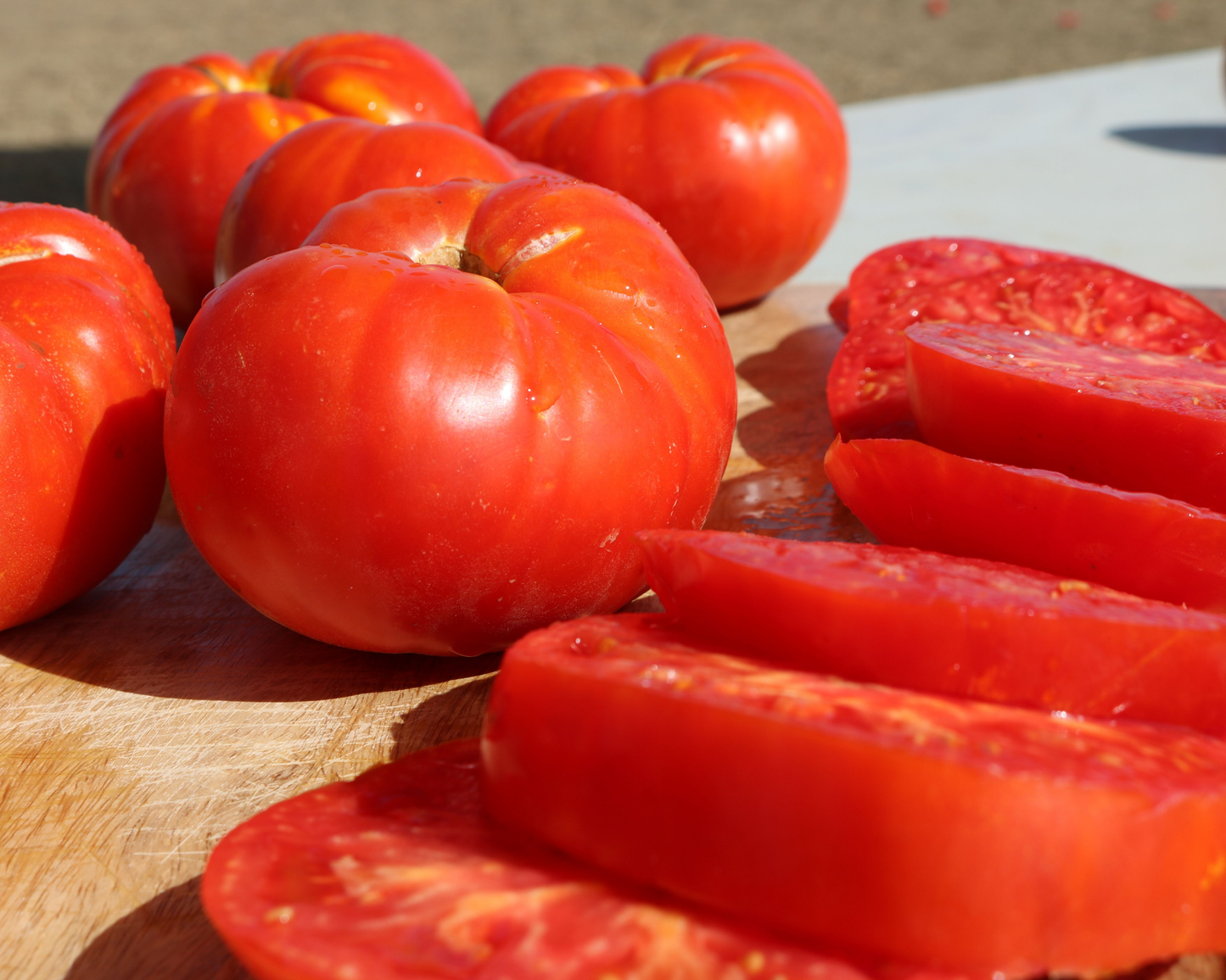 Several whole and sliced Beefmaster Hybrid Tomatoes from Seeds 'n Such are displayed on a wooden cutting board in bright sunlight, highlighting their vibrant red color, juicy texture, and crack-resistant qualities.